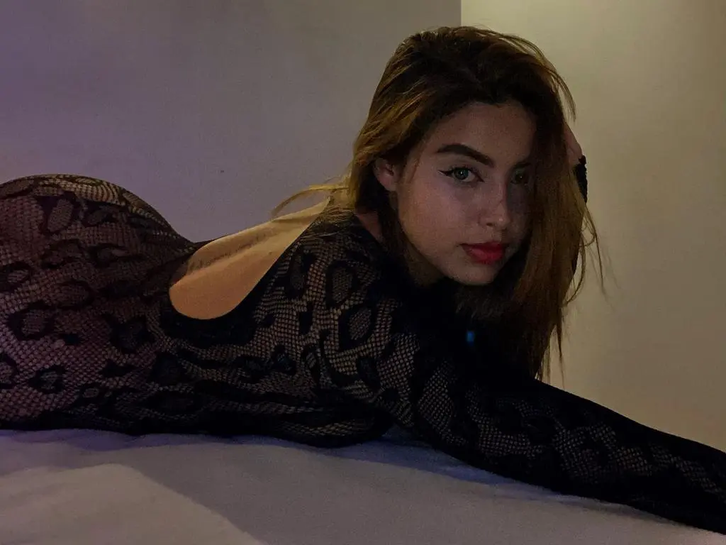 Ariana_Alvarez poses lying down in a black snakeskin print long-sleeve mesh top, looking toward the camera with long brown hair.