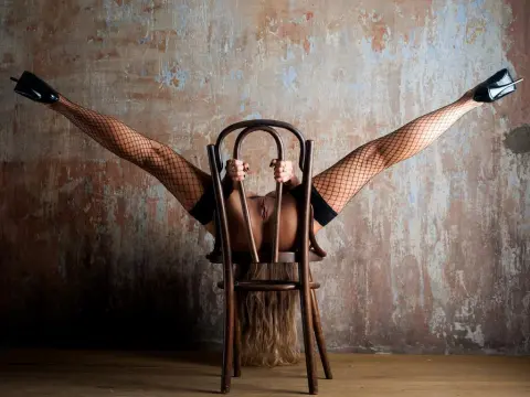 AngelAsPeachmmm — Artistic woman in fishnet stockings and black heels performing a split on a wooden chair against a rustic wall.