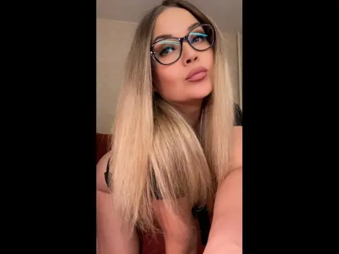 AngelAsPeachmmm — Blonde woman with glasses and long hair posing in a black top for a stylish indoor portrait.