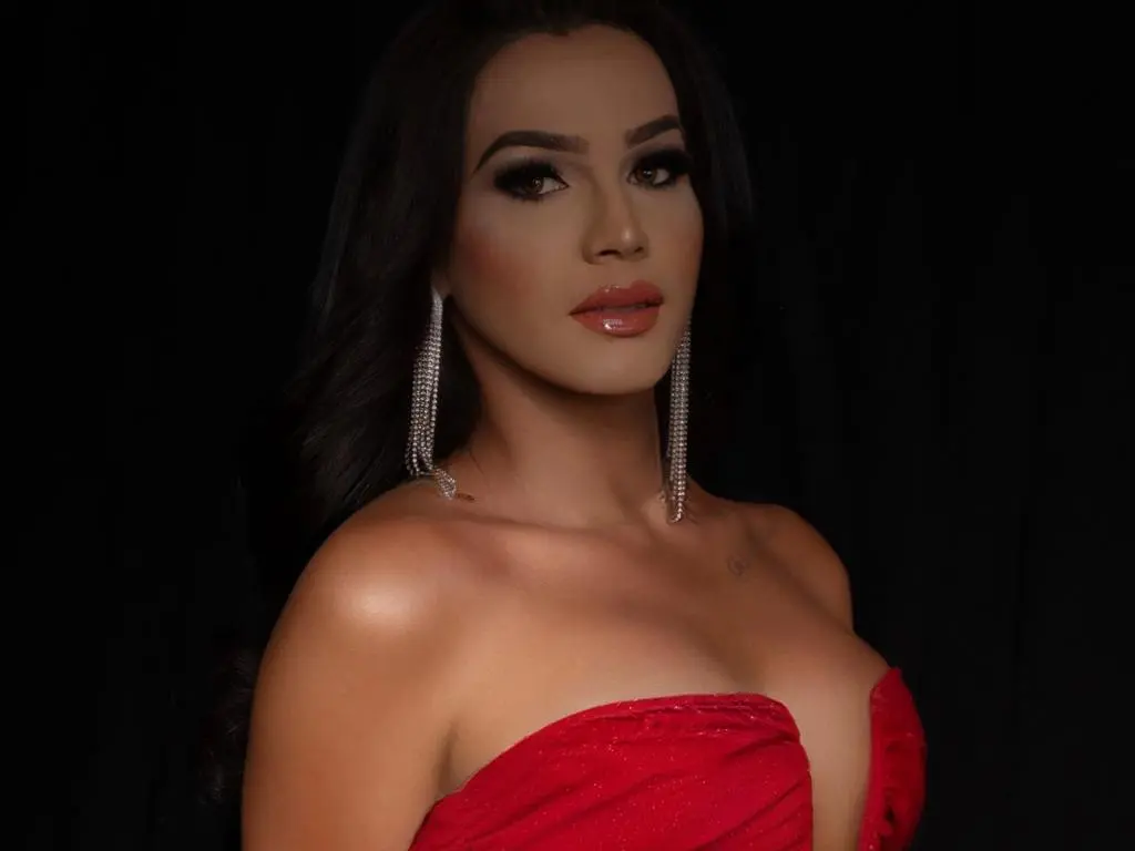 veruzka poses with long dark hair and silver earrings, wearing a red strapless top against a black background.