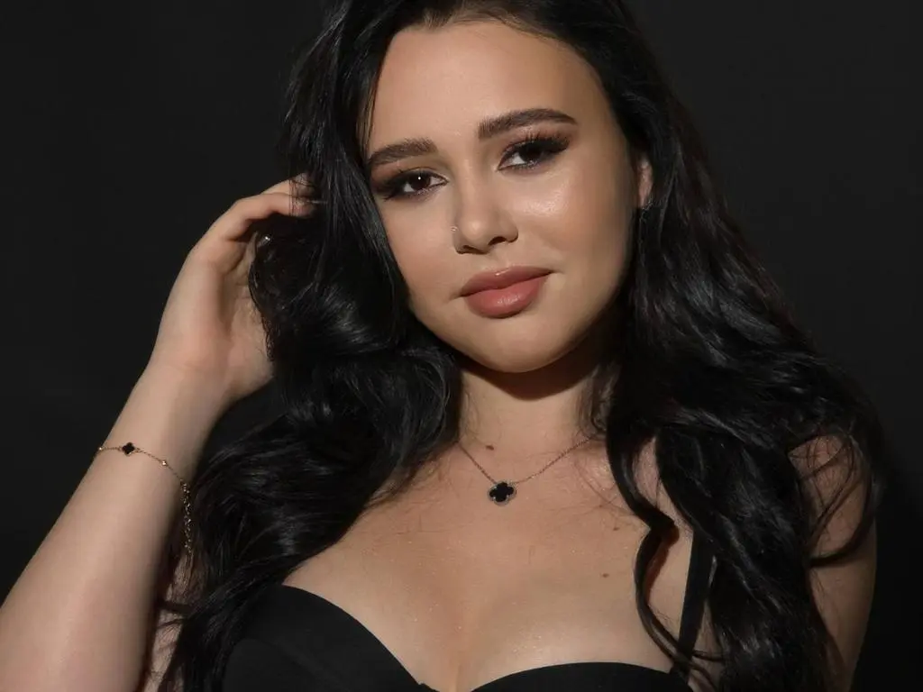 TorryStarPlay poses with her long black hair and a black top against a dark background, looking directly at the camera.