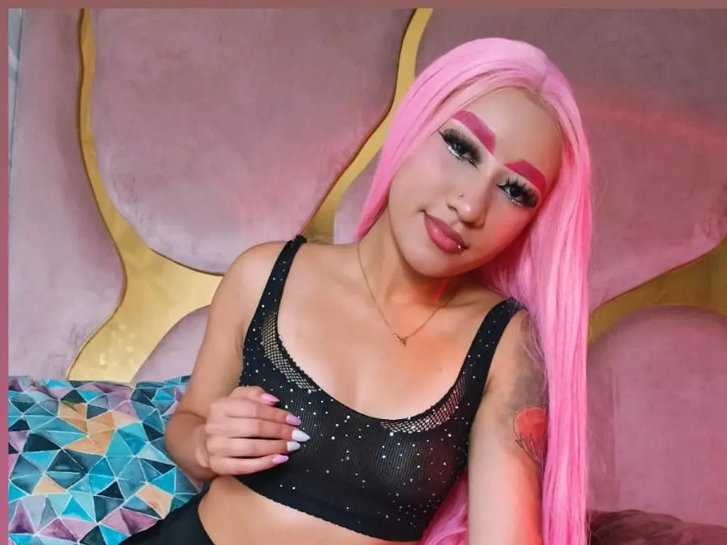 Zafira_Star poses with long pink hair, wearing a black mesh crop top and a gold necklace against a pink headboard.