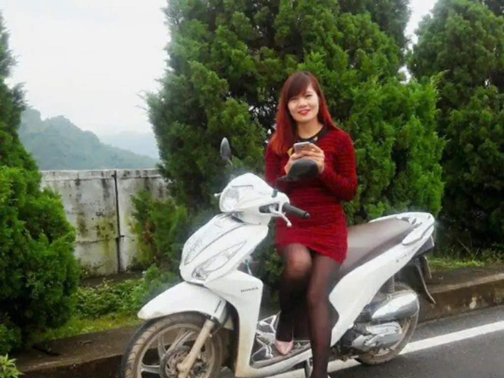 Ellsa93 wearing a red dress and black tights, sitting on a white scooter outdoors against a backdrop of green trees.