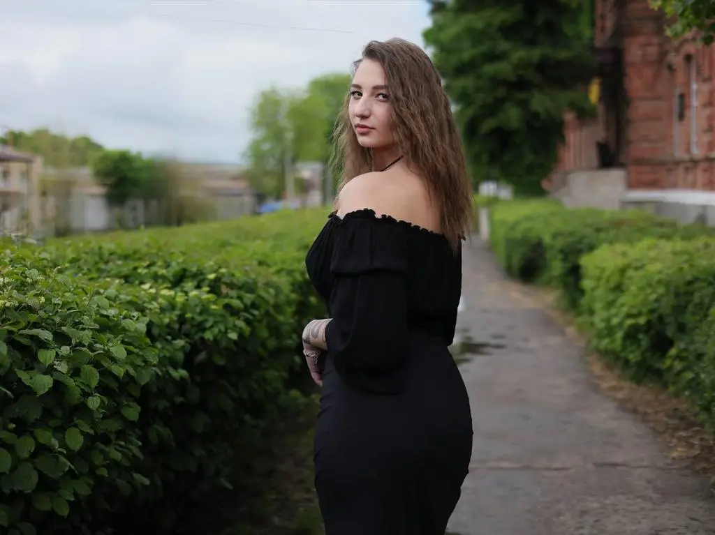 LioraOne poses outdoors in a black off-the-shoulder top and dress, looking back over her shoulder with long brown hair against a background of green hedges.