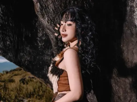 MirannLoves poses against a dark rock wall, wearing a brown furry top and showing her long, wavy black hair and dark eye makeup.