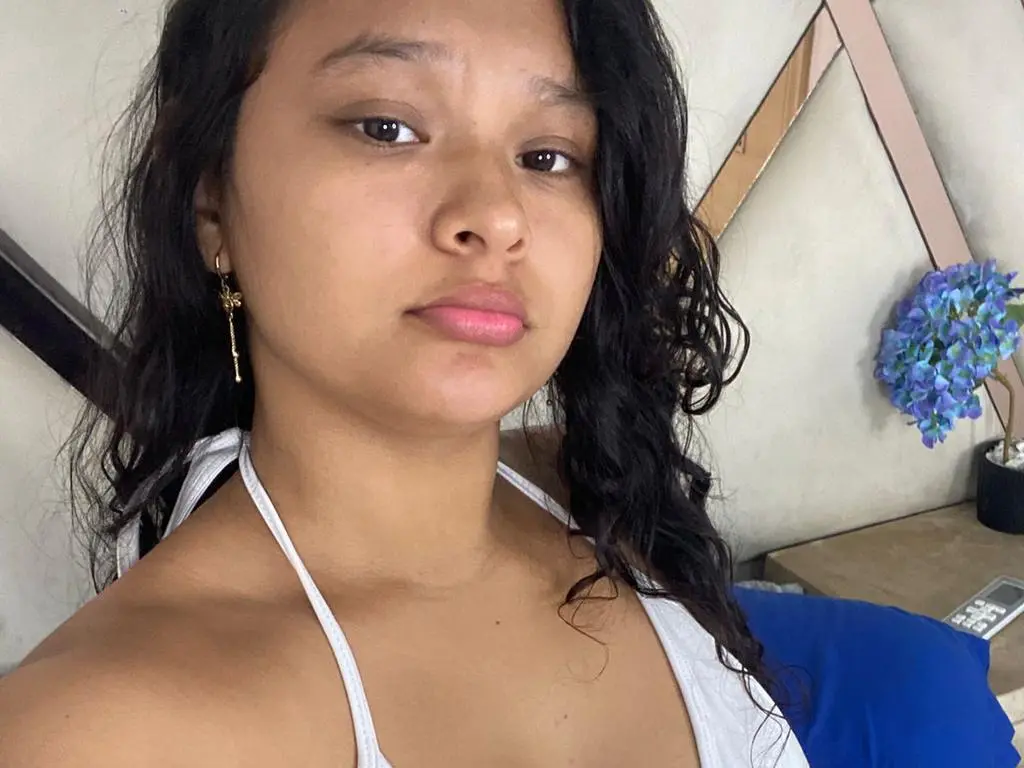 HotJade looks at the camera with long, dark wavy hair and a white halter top, wearing gold earrings in a bright room.