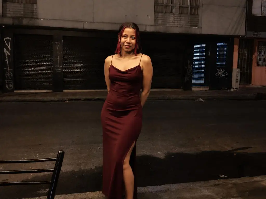 EmilyLovve wearing a long, maroon silk slip dress with thin straps, standing on a dark street at night.