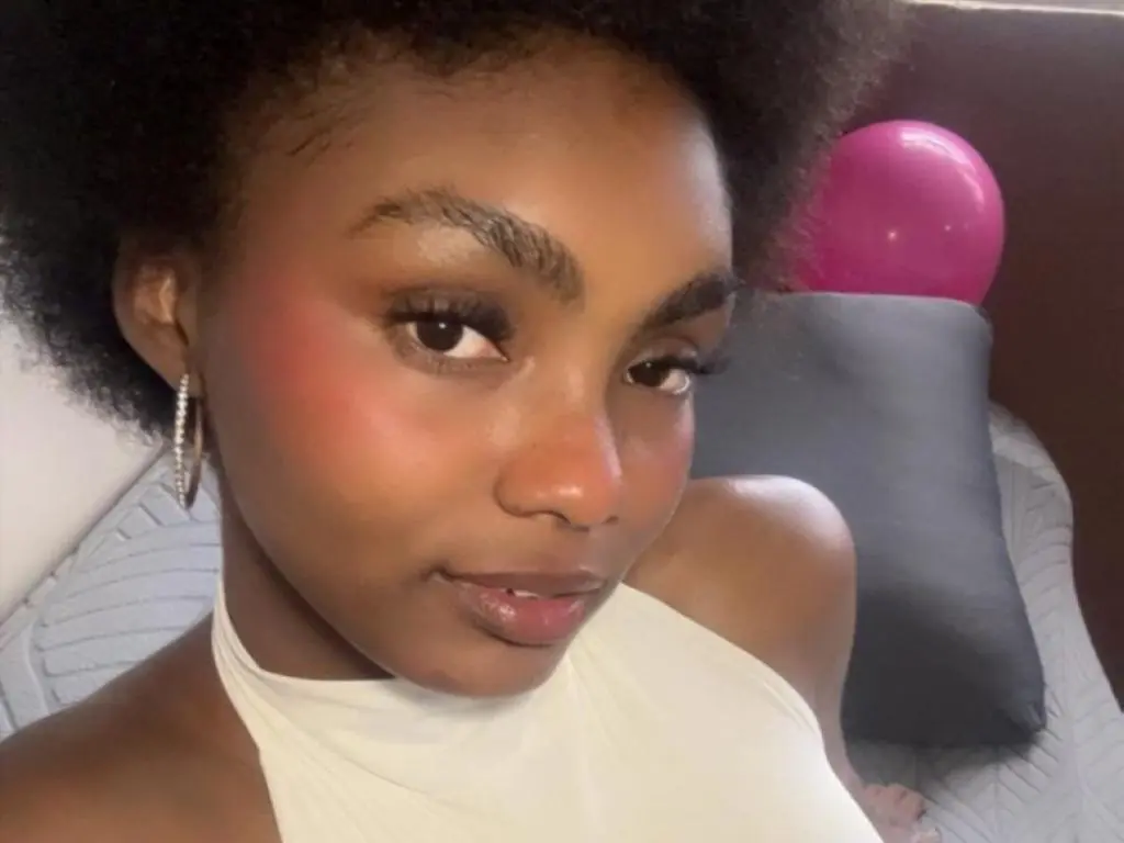 Asha_zuri69 looks directly at the camera wearing a white halter top, showing her face, dark hair, and hoop earrings against a dark background.