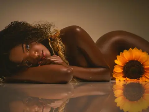 KeisyCollinss — KeisyCollinss lies naked on a reflective surface, posing with her head resting on her arms and her long, curly hair flowing around her near a bright sunflower.