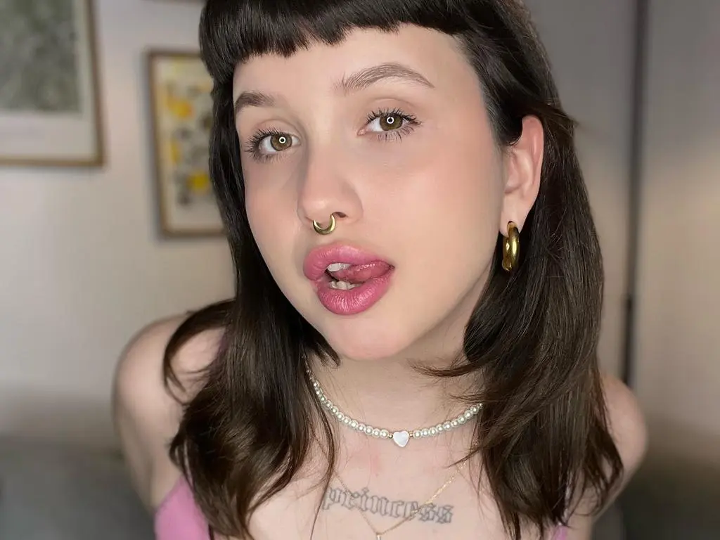 hellfirekiss_ — hellfirekiss_ shows her pink lips and tongue, wearing a pearl choker and gold hoop earrings with a pink top and dark hair with bangs.