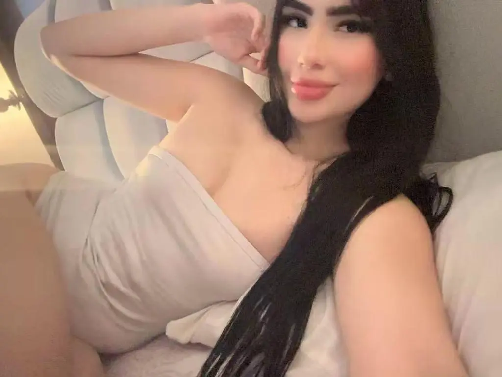 Amber_Rosen lying in bed wearing a white tube top, resting her hand behind her head with her long dark hair falling over her shoulder.