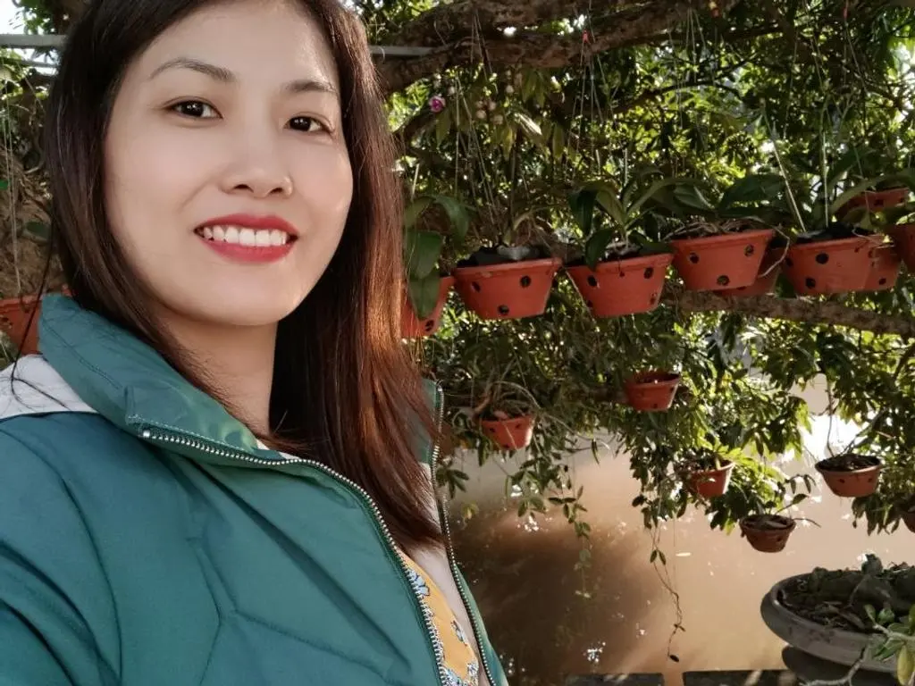 LunaGam smiles outdoors, wearing a green puffer jacket with her long dark hair down, surrounded by hanging potted plants.