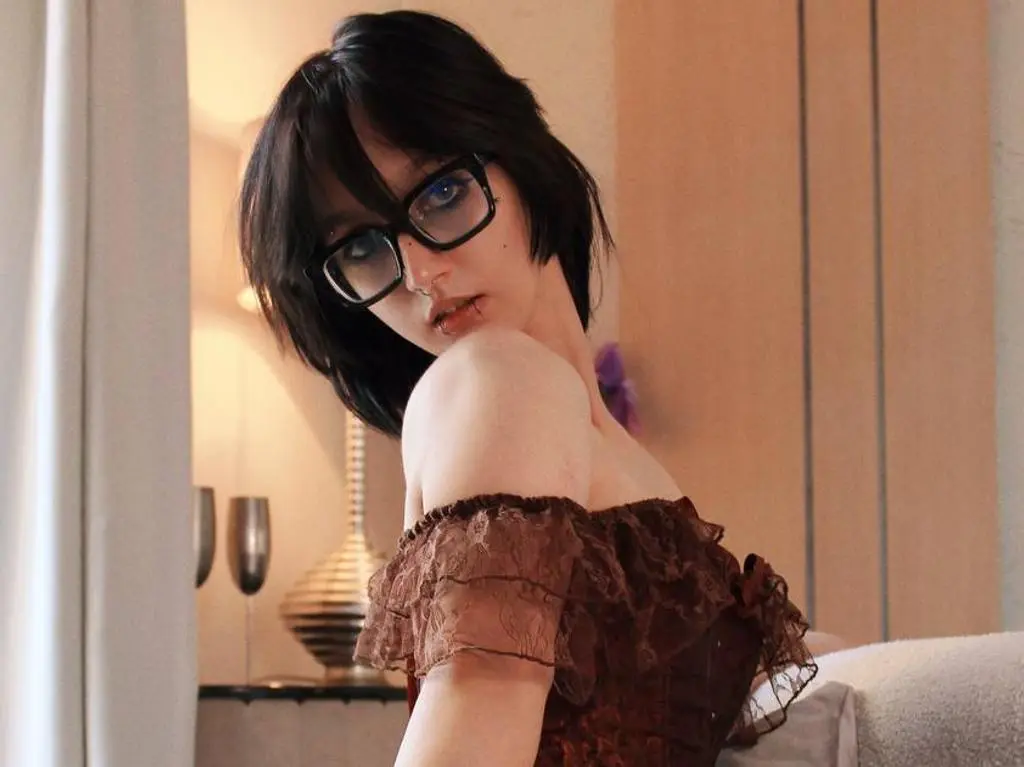 LunaVoguer wearing thick black glasses and a brown lace off-the-shoulder top, looking back over her shoulder with a pouty expression.