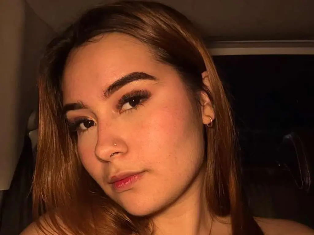 Dani_Suarez poses for a close-up selfie, showing her long brown hair, dark eyebrows, and light makeup in a dim setting.
