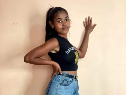 IndianaShowFresh — IndianaShowFresh poses against a plain wall wearing a black cropped tank top and blue denim jeans with a black belt.