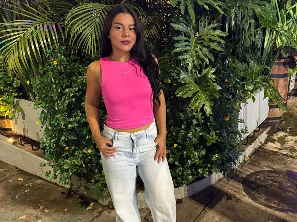 Mia_Bellefort poses outdoors in a pink sleeveless crop top and light blue jeans, standing in front of lush green tropical plants.
