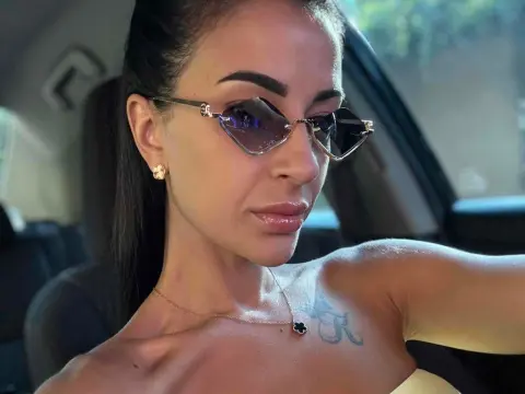 Karinabrunette — Karinabrunette wearing cat-eye sunglasses and a gold necklace, posing in a car with her dark hair pulled back and a tattoo visible on her chest.