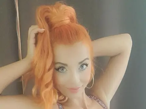 SpicyStarlet — SpicyStarlet poses with her bright orange hair tied in a high ponytail, wearing large hoop earrings and a light-colored bra against a dark wall.