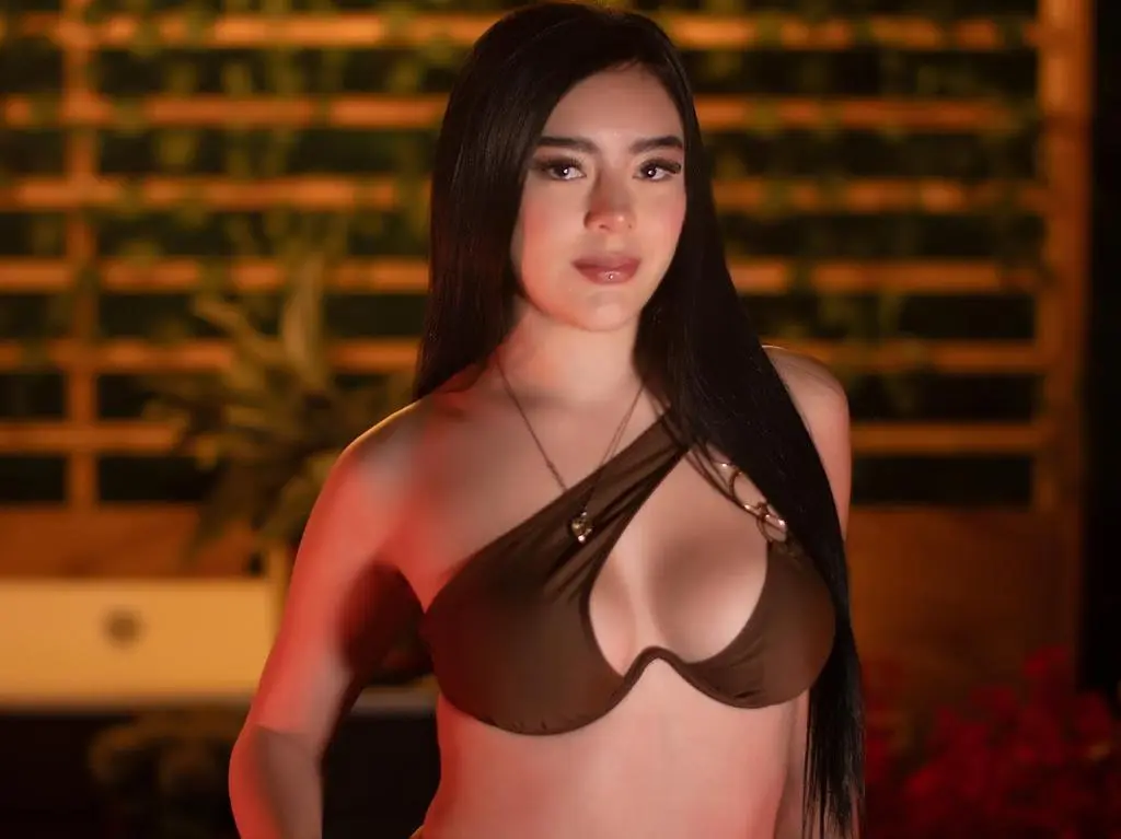 Chanell_Summer poses with long black hair, wearing a brown halter bikini top and a necklace against a dark wooden slatted background.