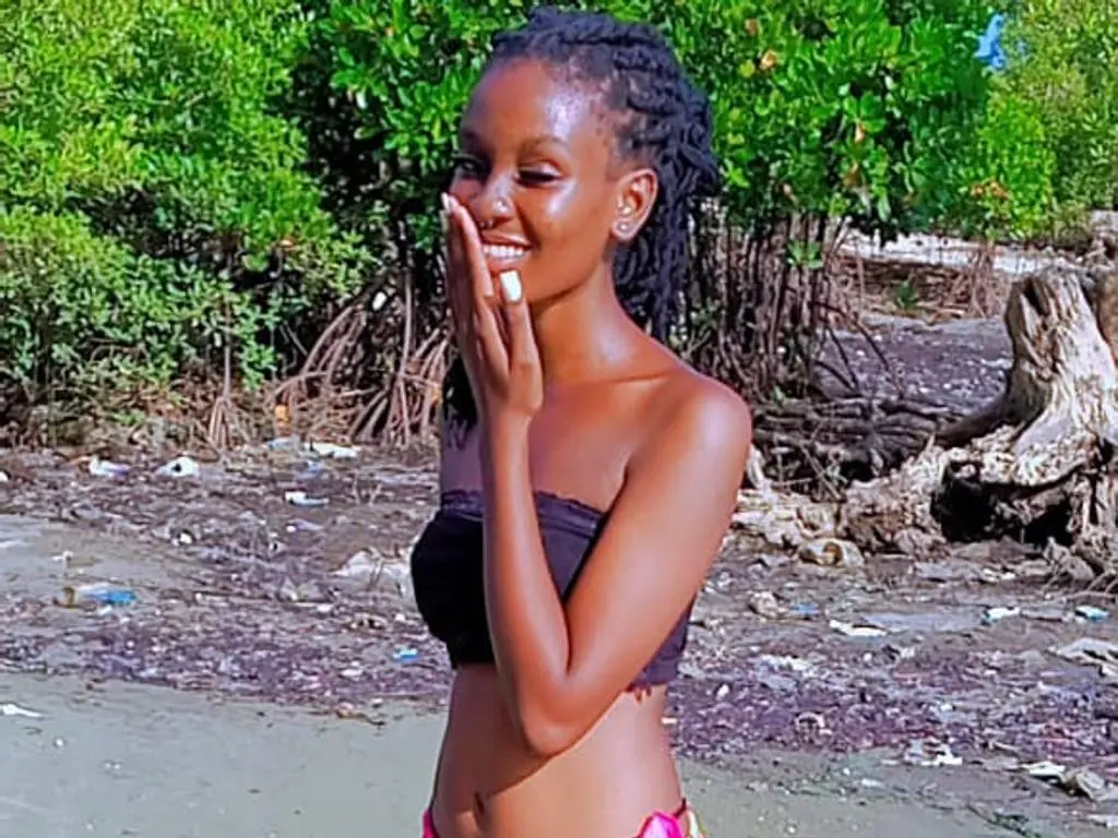 vibesweetershoSmooth smiles while touching her face, wearing a black tube top and pink bikini bottoms in a sunny outdoor setting with green bushes.