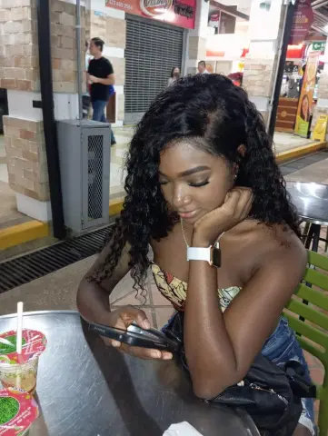 KeisyCollinss — KeisyCollinss with long curly black hair, wearing a strapless yellow patterned top, sitting at a table and looking down at her phone.
