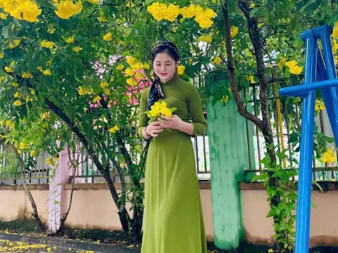JulySex — JulySex wears a long green dress while holding yellow flowers in front of a leafy bush and a blue metal frame.