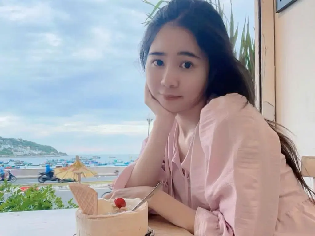 Emily2011 with long dark hair wearing a pink shirt, resting her chin on her hand while sitting at a table with a dessert outdoors.