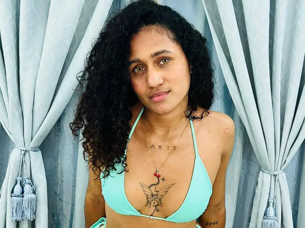 Kynesa is a curly-haired woman wearing a light blue bikini top and a gold necklace, posing in front of light blue curtains.