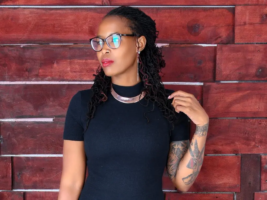 Ceciliaxxx wears glasses and a black t-shirt with a copper neck ring, posing against a dark wood wall with her long dreadlocks and arm tattoos visible.