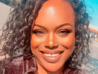 1lookurhooked smiles with her long, dark curly hair framing her face in a close-up headshot.