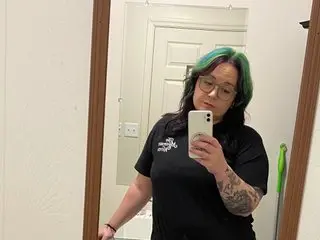 5lut_puppy poses in a black t-shirt, looking into a mirror with her dark hair styled with green highlights in a bright room.