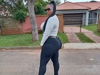 AFRICANCURVYBOOTYXXX poses sideways in a white long-sleeve top and tight black jeans, showing off her curves against a background of trees and a house.