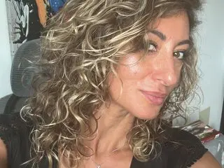 AKALYI poses with her long, curly blonde hair, showing her bare shoulders and chest in a close-up shot.