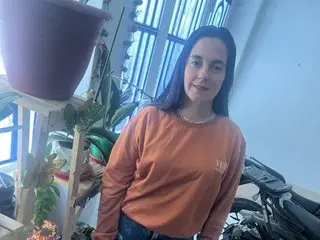 Smiling woman in an orange long-sleeve shirt standing near indoor plants in a bright room.