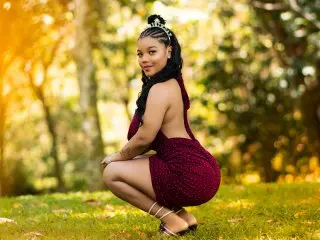 ASHAANTI is a beautiful Black woman with long braids, wearing a tight red dress while posing in a sunny park.