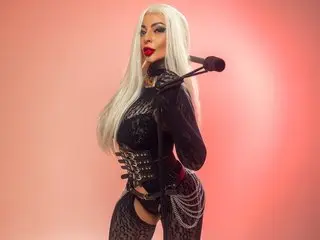 AbbyPink69 poses in a black lace bodysuit with long white hair and red lipstick against a pink background.