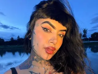 Portrait of a tattooed woman with dark hair and winged eyeliner posing outdoors during a blue twilight sunset.