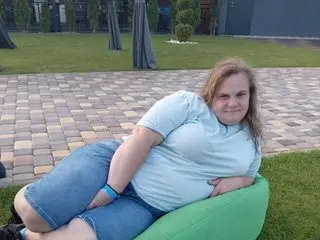 Adela_Cheri wearing a light blue t-shirt and denim shorts, sitting on a green beanbag chair outdoors on a paved patio.
