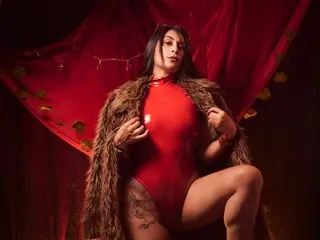 AfroditaDior poses in a red bodysuit and fur coat against a red curtain backdrop.