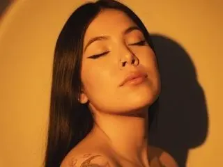 AilynRed poses with her long dark hair and eyes closed, showing her bare shoulders and chest against a warm yellow background.