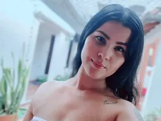 AishaAmin looks at the camera with long black hair and bare shoulders in an outdoor setting.