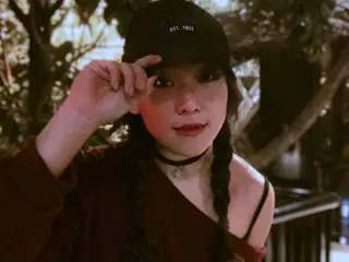 Aishapark looks at the camera with her hair in two long braids, wearing a black crop top and a black choker against a soft outdoor background.