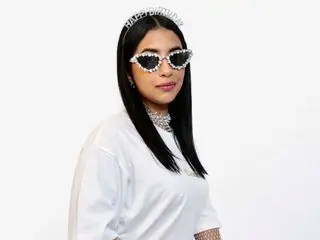 Alana_mitchell wears a white t-shirt, sparkly sunglasses, and a birthday tiara while posing against a white background.