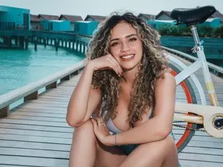 AleCollins smiles while sitting on a wooden pier next to a bicycle, wearing a blue bikini top and showing off her long curly hair.