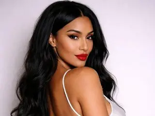 AleciaX poses with long, wavy black hair and red lipstick, wearing a white strap against a plain light background.