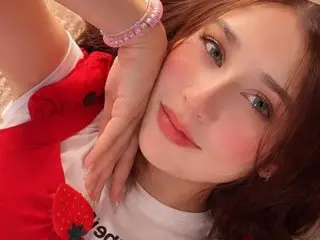 Smiling young woman in a red strawberry outfit and pink bracelet posing for a bright, cheerful close-up selfie.