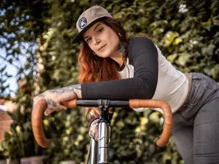 AlessaMoth poses outdoors with her hands on the handlebars of a bicycle, wearing a tan cap and a black and white long-sleeved top.