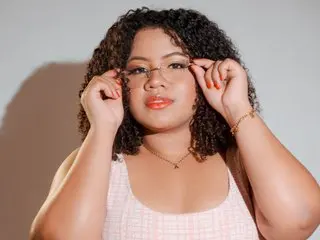 Alessia_kaun looking at the camera with her hands touching her glasses, wearing a white tank top with her dark curly hair down.
