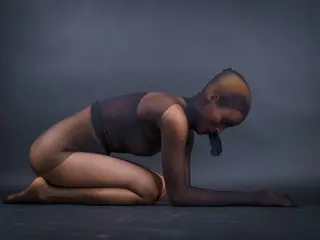 AlexaDuque kneels on a dark surface, wearing a black bra and posing with her head down, showing her bare back and legs.
