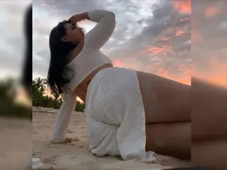 Curvy woman in white summer outfit posing on a tropical beach during a dreamy pink sunset.